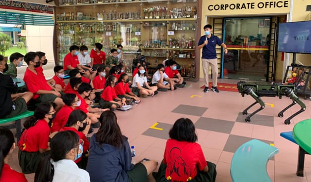 A presenter speaks to masked students seated on the floor near a four-legged robot dog in a corporate office area.